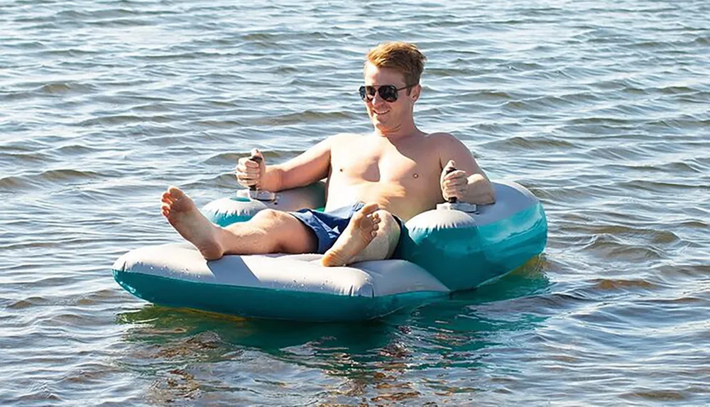 A man in sunglasses is relaxing on a floating inflatable armchair on the water giving a thumbs-up with both hands