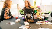 Two people are seated at a table with festive decorations and a sign suggesting a fortune-telling theme.