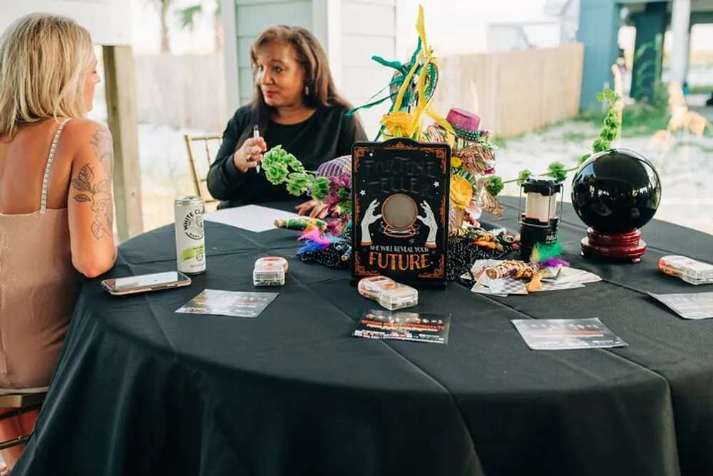 A woman is sitting at a table adorned with mystical and festive decorations possibly engaged in a fortune-telling session with another person whose back is to the camera