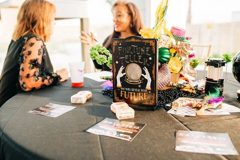 Two people are conversing at a table adorned with a festive Fortune Teller sign and party decorations suggestive of a themed event