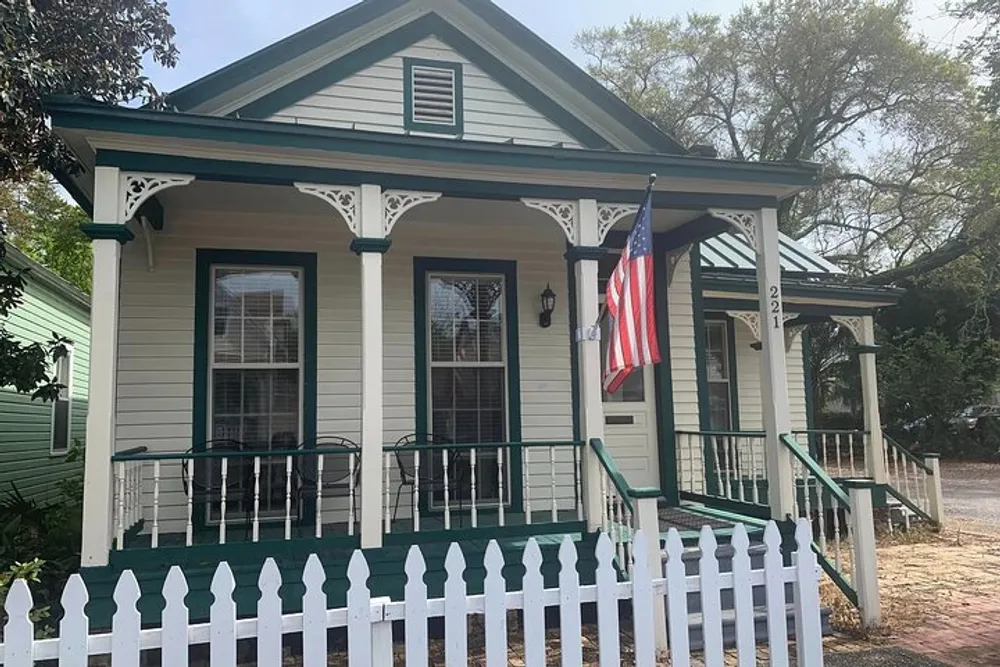 The image displays a charming traditional American house with a welcoming front porch green trim and an American flag