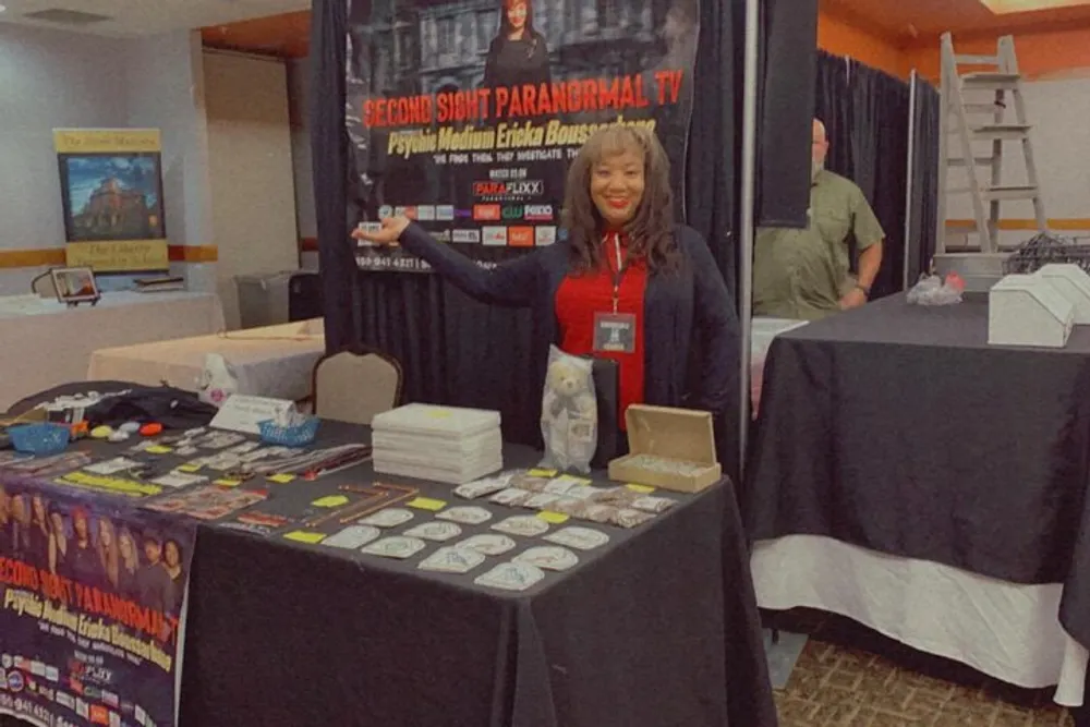 A smiling person is standing behind a vendor booth displaying books flyers and merchandise related to Second Sight Paranormal TV