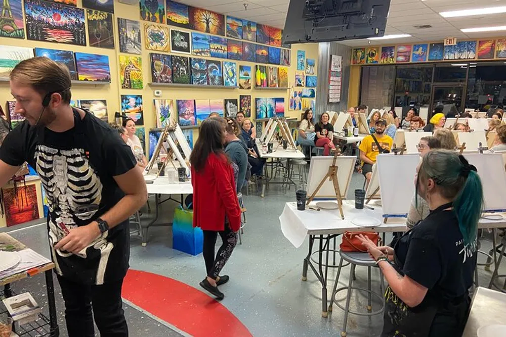 A group of people is gathered in a vibrant art studio engaging in a painting class with easels set up and colorful artwork adorning the walls