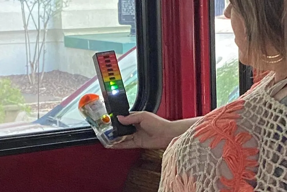 A person is holding a device with color-coded lights while sitting near a window possibly on a bus or train