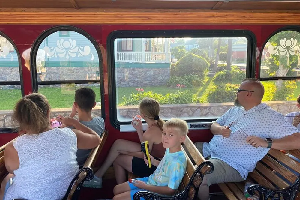 Passengers including a young boy looking at the camera are seated inside an old-fashioned trolley car with large windows enjoying a sunny day