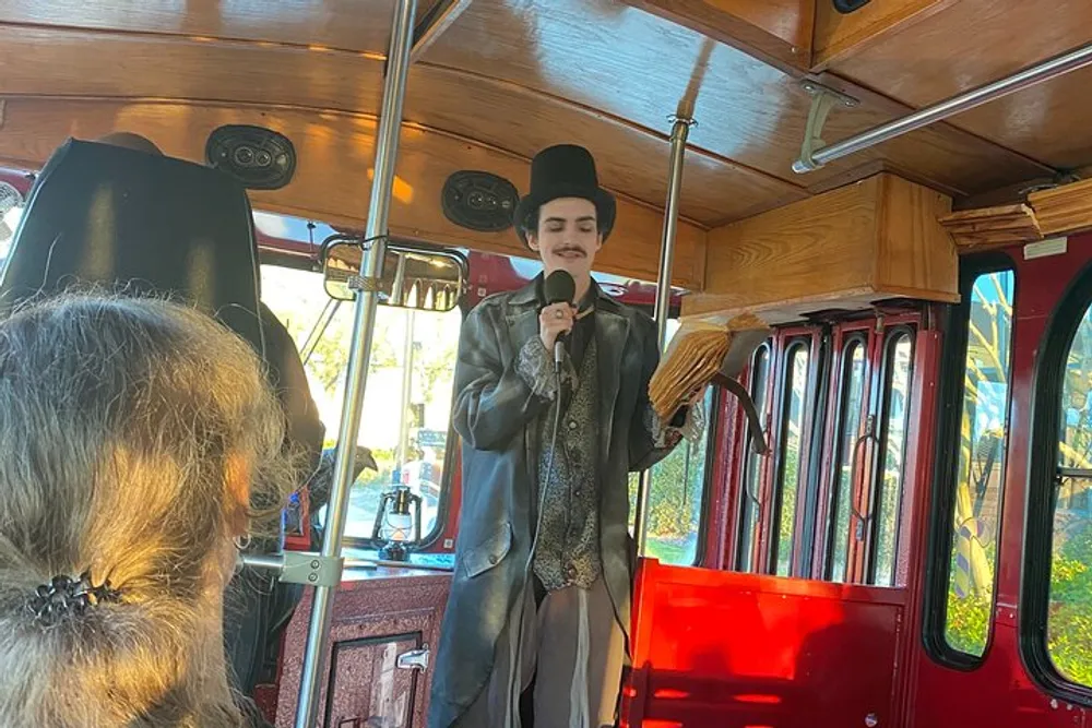 A person dressed in vintage-style clothing including a top hat is standing and speaking into a microphone onboard a trolley or vintage-style bus with wooden features and red trim