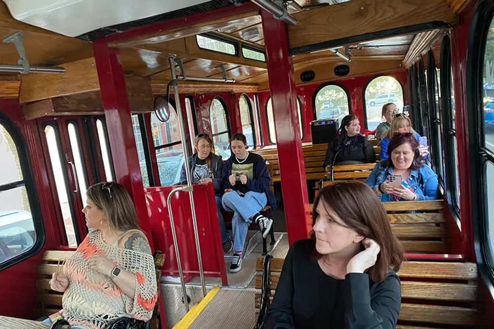 Passengers are seated inside a vintage-style trolley or streetcar with wooden benches and red trim some engaged with their phones while others look around or converse