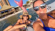 Two people are smiling for a selfie while floating on inflatables in the water with a beachfront building in the background.