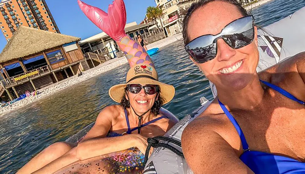 Two people are smiling for a selfie while floating on inflatables in the water with a beachfront building in the background