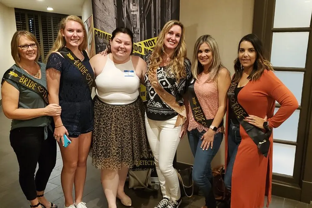 A group of women is smiling for the camera at what appears to be a pre-wedding celebration with some wearing sashes that read Bride Tribe and Bride set against a backdrop featuring Crime Scene Do Not Cross tape