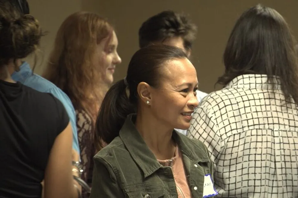 A group of people are engaged in a relaxed social interaction with a smiling woman in the foreground wearing a jacket and a nametag