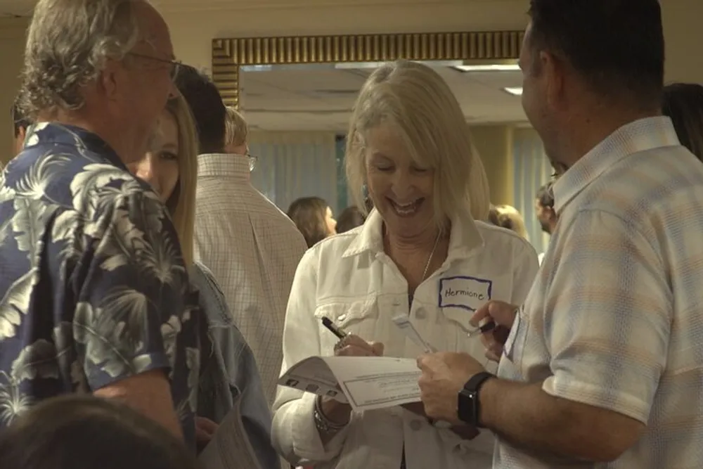 A group of adults are interacting and smiling with one woman wearing a name tag and reading from a paper at what appears to be a social or professional event