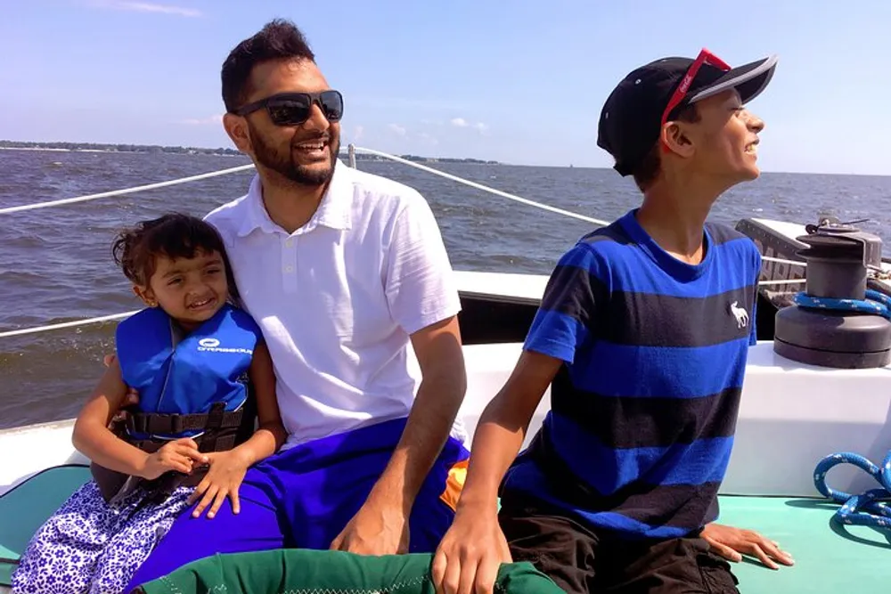 A man and two children are enjoying a sunny day out on a boat with the man smiling at the camera a young girl in a life jacket snuggling up to him and a boy looking away into the distance with interest