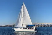 A sailboat with its sails fully unfurled glides through the water near a long bridge under a clear blue sky.