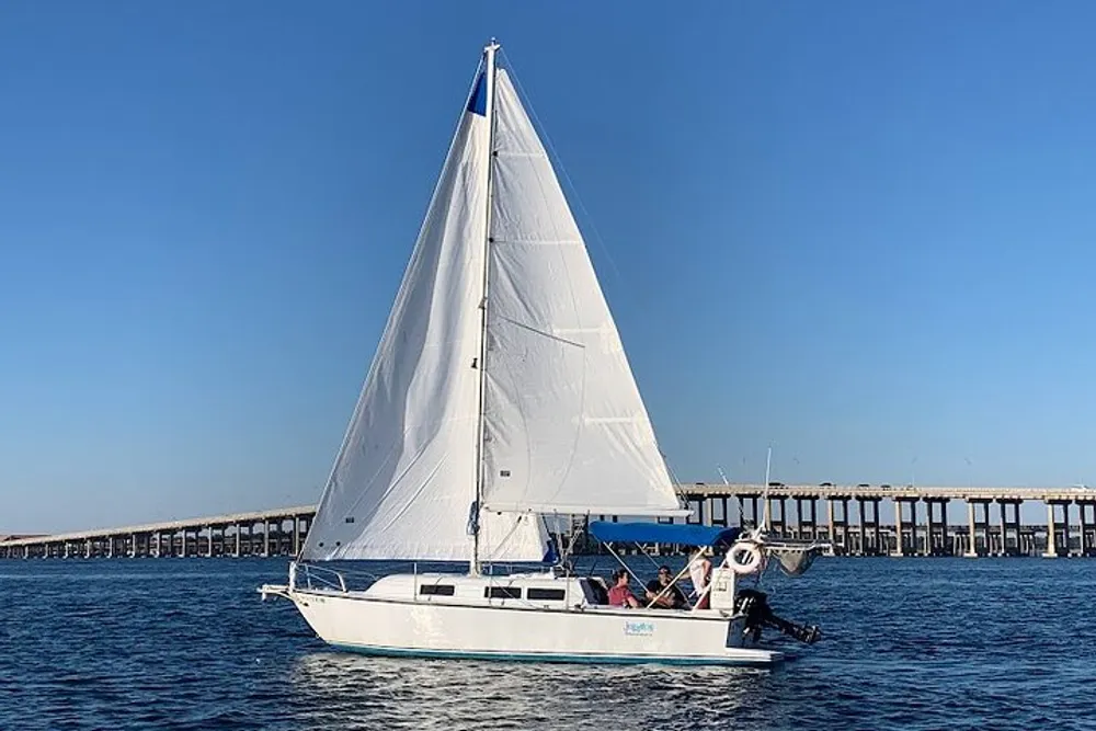 A sailboat with its sails fully unfurled glides through the water near a long bridge under a clear blue sky