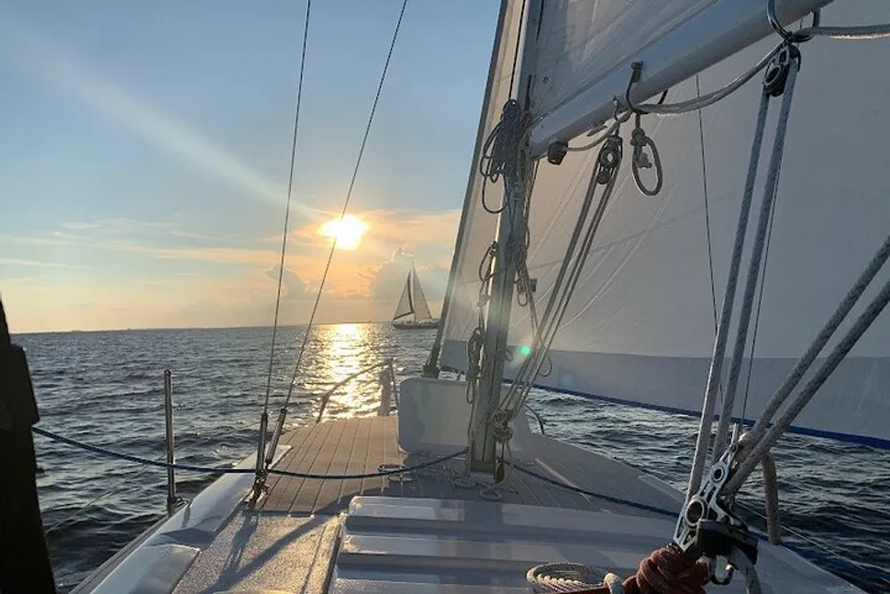 The image captures a serene sailboat experience with the setting sun and another sailboat in the distance as viewed from the deck of a sailboat