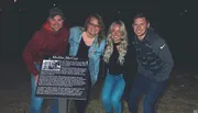 Four individuals are smiling for a photo at night, with one of them holding a sign about a historical figure named Mollie McCoy.