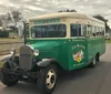 A vintage-style green and cream Hop On Hop Off tour bus is parked on a sunny tree-lined street with historic houses in the background