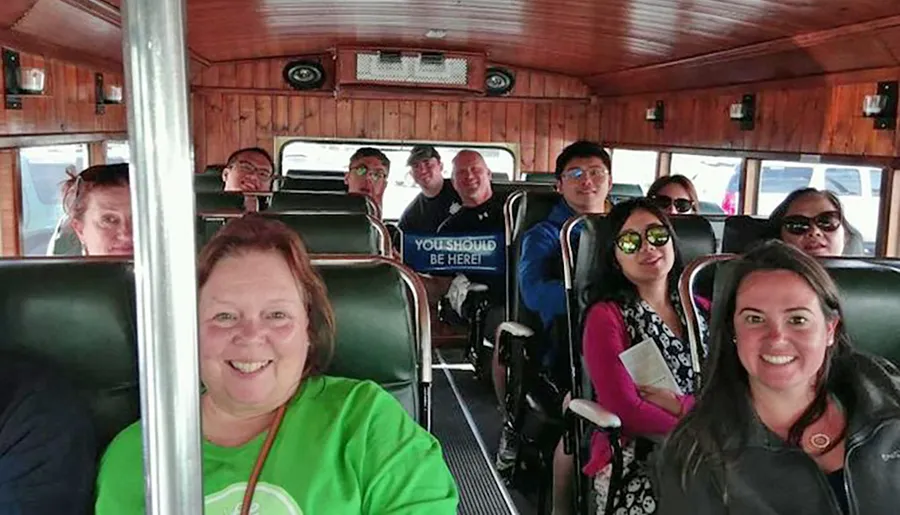 A group of smiling people are seated inside a vehicle with wooden interior, posing for a photo, with one person holding a sign that says YOU SHOULD BE HERE.