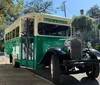 A vintage-style green and cream Hop On Hop Off tour bus is parked on a sunny tree-lined street with historic houses in the background