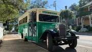 A vintage-style green and cream 'Hop On Hop Off' tour bus is parked on a sunny, tree-lined street with historic houses in the background.
