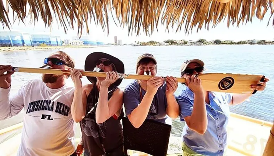 Four people are humorously pretending to bite into a long wooden paddle together under a thatched roof, possibly on a boat, with a body of water and shoreline in the background.