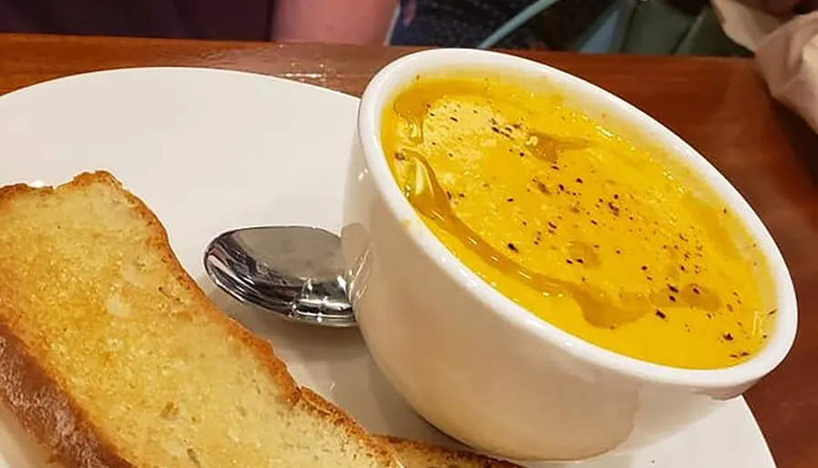 A bowl of soup garnished with black pepper is served next to a slice of toasted bread and a spoon on a white plate.