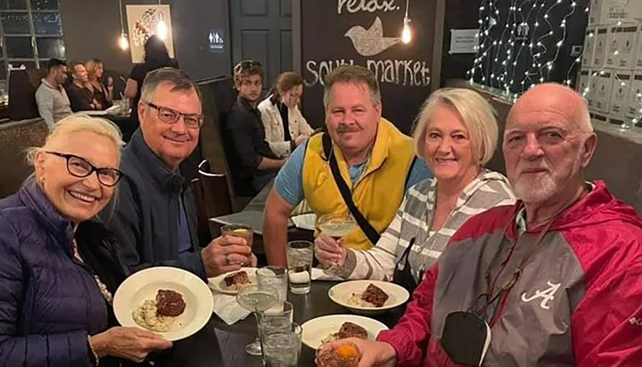 A group of five smiling adults is gathered around a restaurant table with drinks and plates of food in a cozy dining setting.