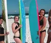 A person in a tie-dye shirt is posing on a surfboard on the sand with two children doing the same behind them under a bright sun with a beach umbrella and various beach items around