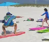 A person in a tie-dye shirt is posing on a surfboard on the sand with two children doing the same behind them under a bright sun with a beach umbrella and various beach items around