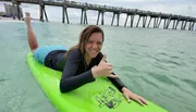 A smiling person is giving a thumbs-up while lying on a green surfboard in the water near a pier.