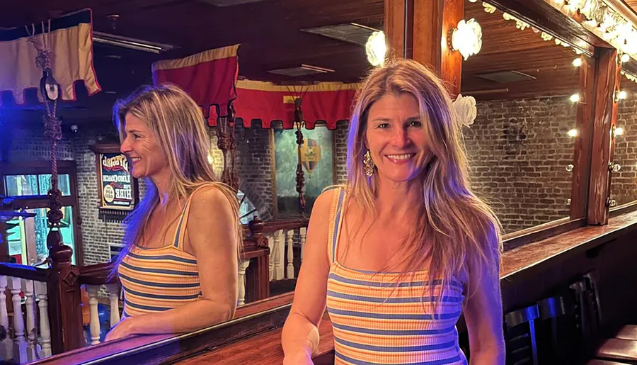 A smiling woman in a striped tank top stands at a polished wooden bar with a mirrored backdrop reflecting her image.