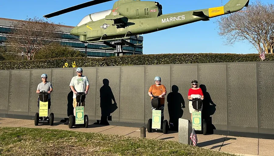 Four people wearing helmets are standing on Segways in front of a wall memorial with a military helicopter on a pedestal in the background.