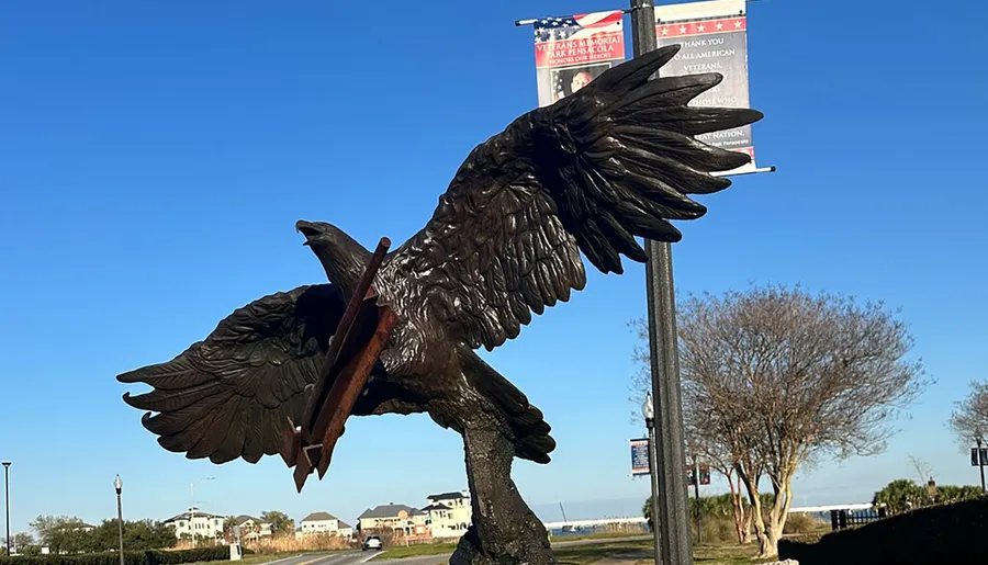 The image shows an intricately detailed sculpture of an eagle with outstretched wings, mounted on a base, with a clear blue sky in the background and a banner honoring veterans visible above it.