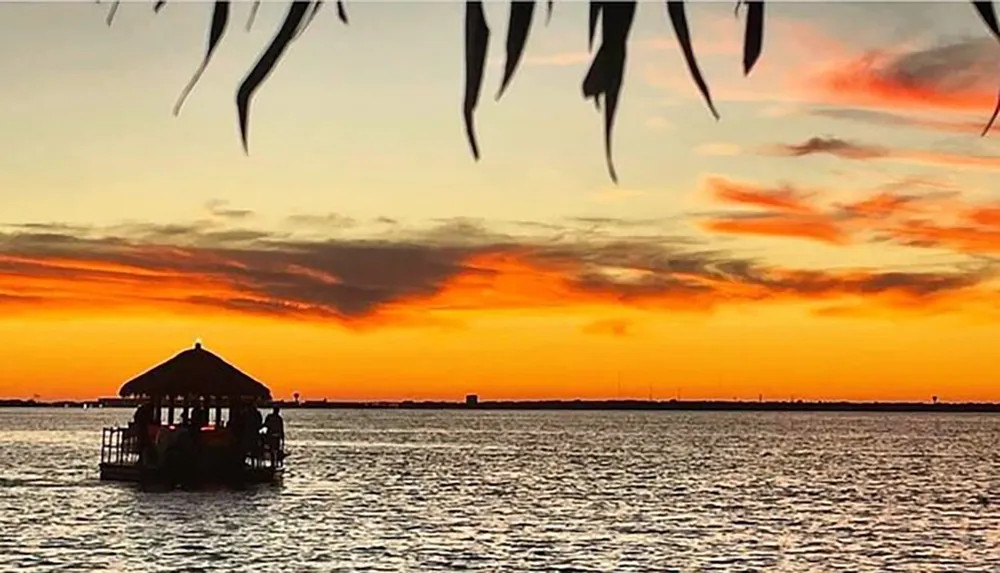 A thatched-roof structure floats on calm waters under a vibrant sunset sky with the silhouette of palm leaves framing the top of the image