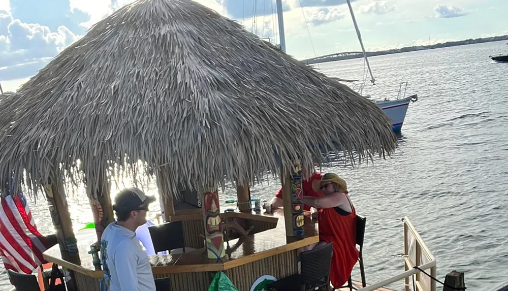 People are enjoying a sunny day at a tiki bar by the water with a sailboat in the background