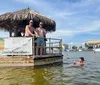 Two individuals are standing on a floating tiki bar structure while another person is swimming in the water next to them all seemingly enjoying a sunny day on the water