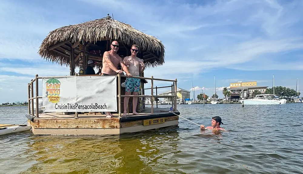 Two individuals are standing on a floating tiki bar structure while another person is swimming in the water next to them all seemingly enjoying a sunny day on the water