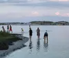 A group of people including children and adults are walking along a narrow strip of land that stretches into the water reflecting the cloudy sky above at dusk