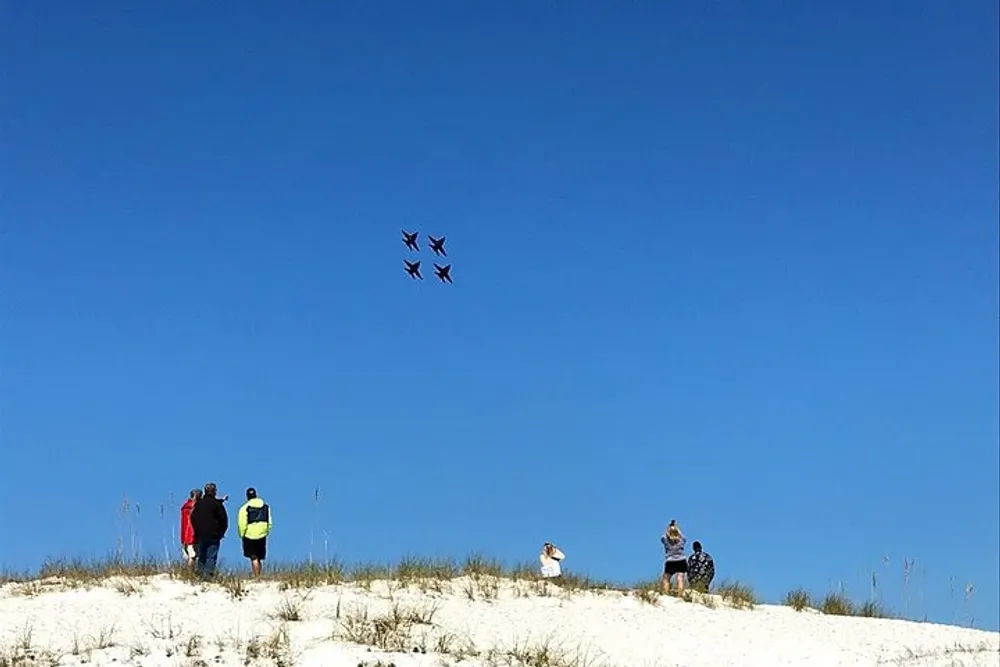 A group of people standing on a sandy hill are watching a formation of jets flying overhead against a clear blue sky