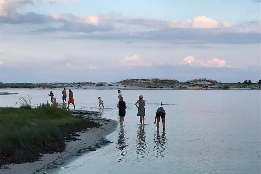 Several people are wading and walking along the edge of a tranquil body of water with a sky of pastel hues as the backdrop