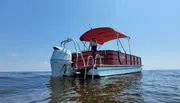 A person is standing on a pontoon boat with a red bimini top, anchored in calm waters under a clear blue sky.