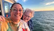 Two people are smiling for a selfie with a serene ocean backdrop and a bridge on the horizon during a sunset.