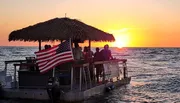 A group of people enjoys a scenic sunset from a thatched-roof pontoon boat flying the American flag on the ocean.
