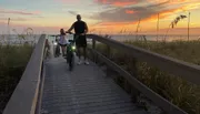 Two people on bicycles are enjoying a scenic sunset by the beach on a wooden boardwalk.