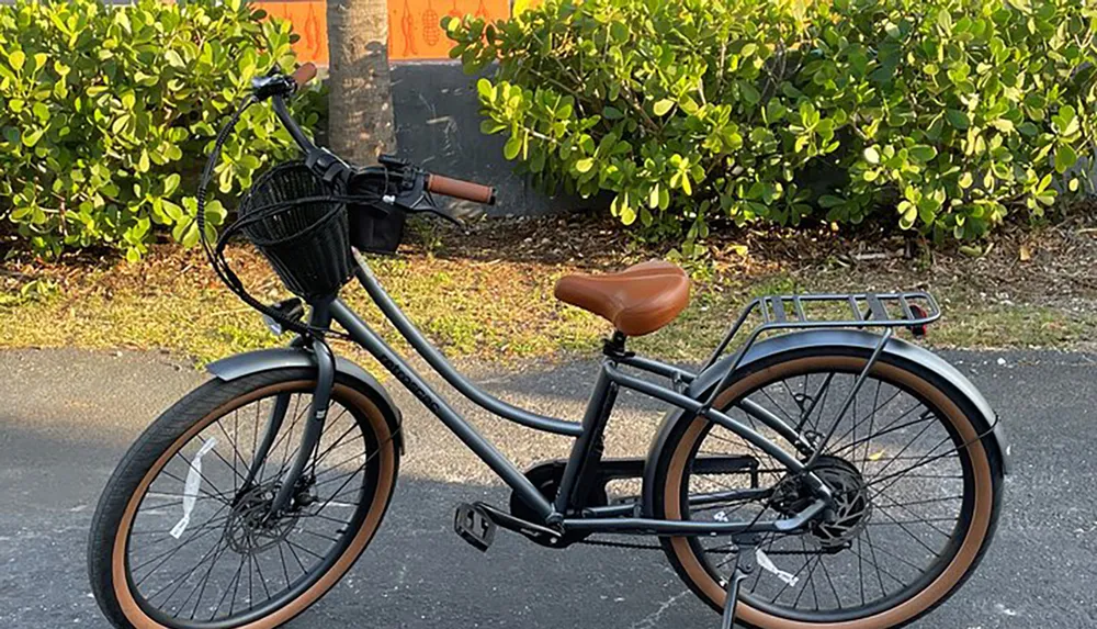 A modern black bicycle with a brown saddle and grips and a front basket is parked on a pavement beside a lush green hedge