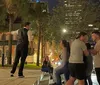 A man in a vest and white shirt is performing or demonstrating something at night to an attentive group of onlookers in an urban palm-lined setting
