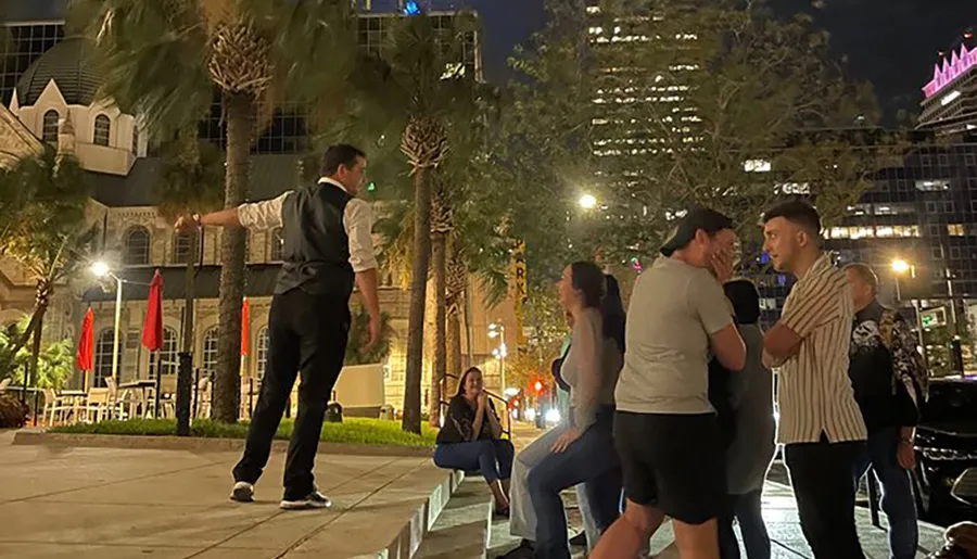 A man in a vest and white shirt is performing or demonstrating something at night to an attentive group of onlookers in an urban, palm-lined setting.