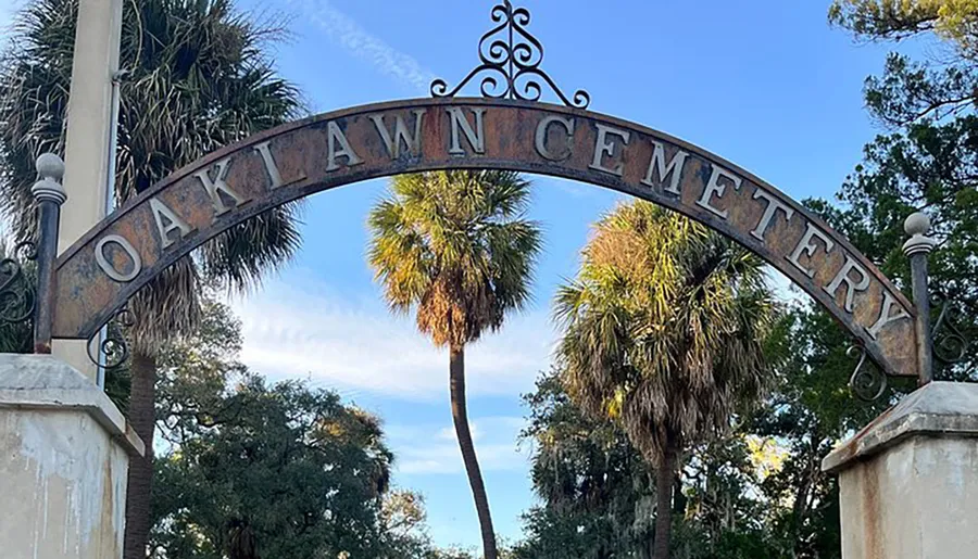 The image shows an arched metal sign with the inscription OAKLAWN CEMETERY framed by a blue sky and flanked by palm trees.