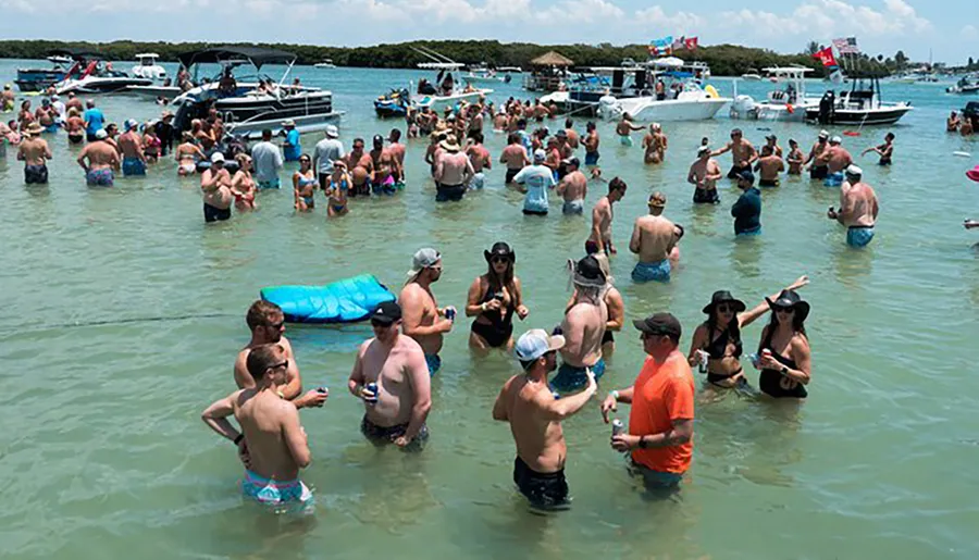 A group of people is socializing in shallow waters near several boats, indicating a water-based gathering or event.